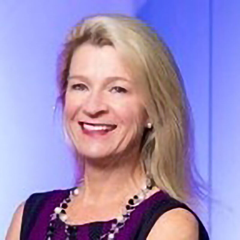 The image shows a woman with blonde hair, smiling at the camera. She is wearing a dark top with a purple and black pattern and a beaded necklace. The background is a blurry blue.